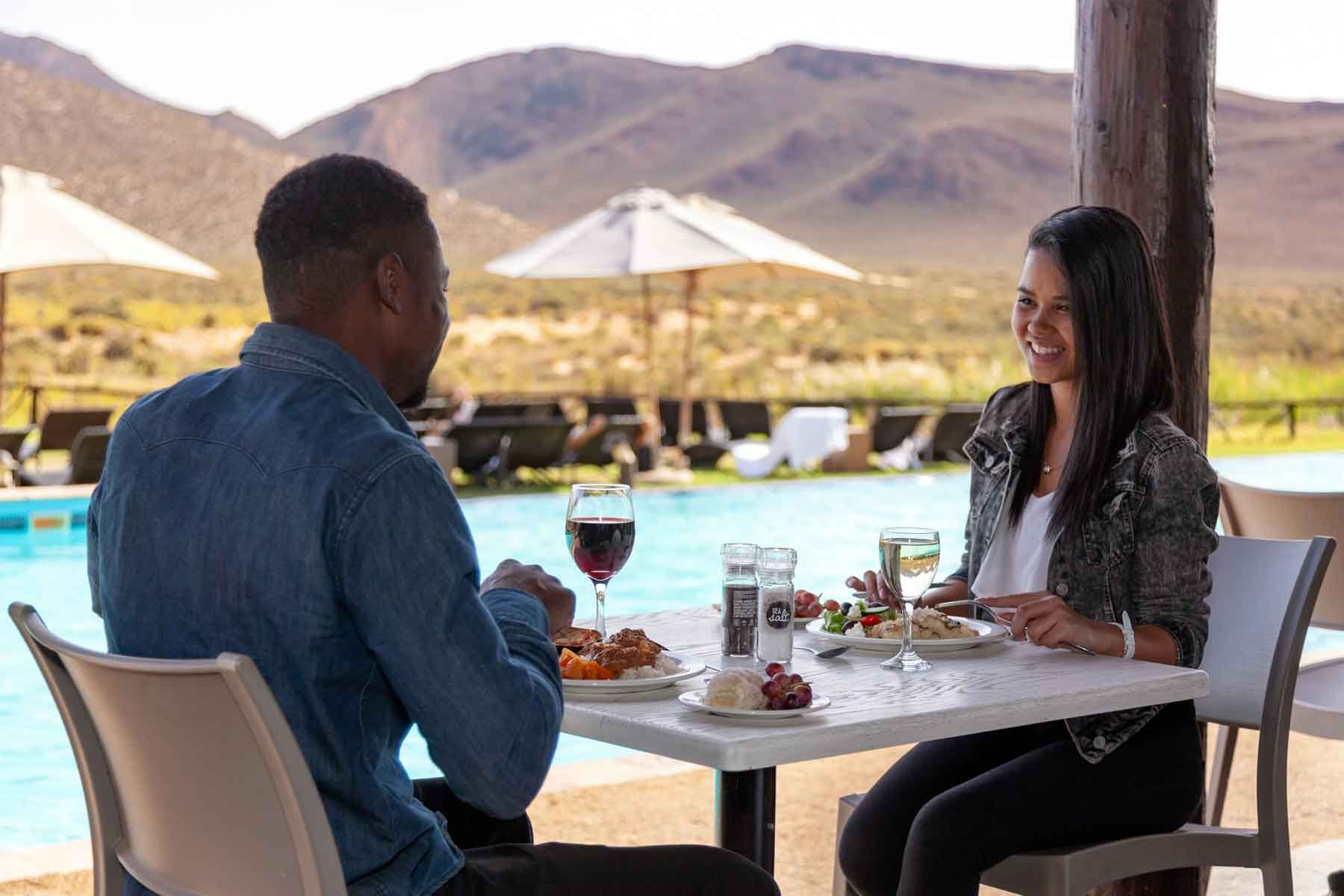 MoztoCape Guests dining poolside, mountains in the background. Woman smiles, man eats. Table with food, wine glasses.