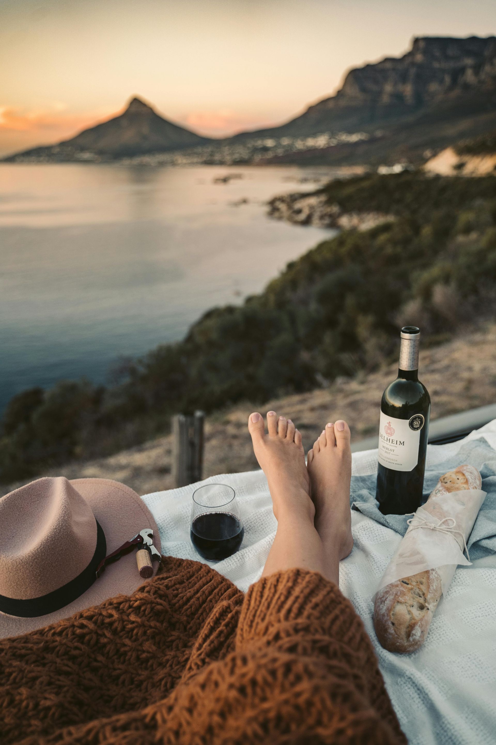 Person relaxing with wine and bread, scenic seaside view, hat and sweater.