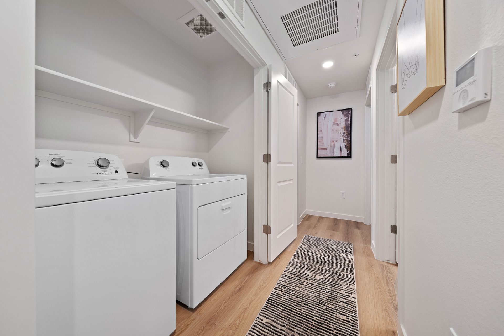 Laundry room with white appliances, shelves, and a patterned rug on wooden flooring.