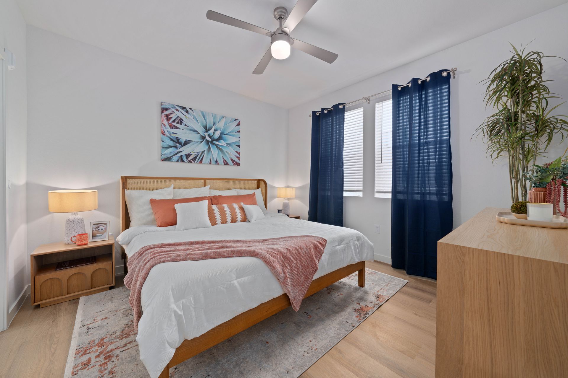 Bedroom with wooden bed, white bedding, pink accents, blue curtains, and a ceiling fan.