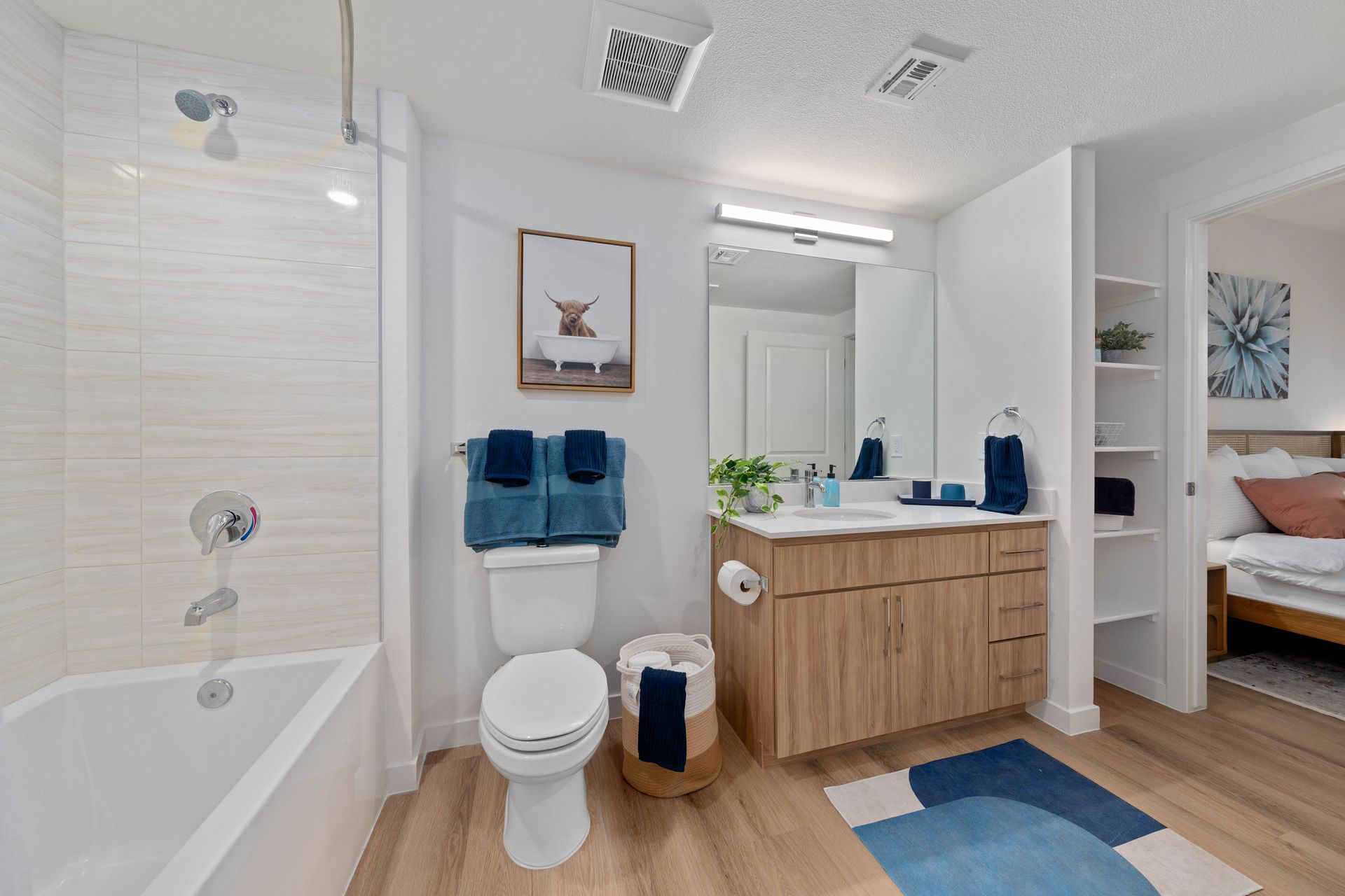 Bathroom with a tub, toilet, sink, and a view into a bedroom. White, blue, and light wood tones.
