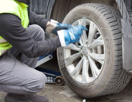 Person changing a tire on a gray car, wearing a yellow vest and blue gloves.