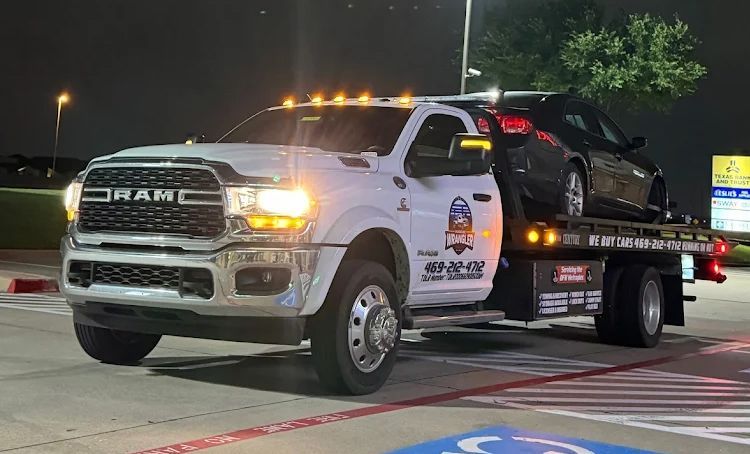 White tow truck hauling a black car at night. Truck has flashing amber lights.
