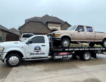 Tow truck with tan pickup truck on its bed, in front of a brick house on a cloudy day.