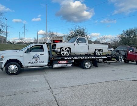 White tow truck towing a classic white pickup truck and a red truck under a blue sky.
