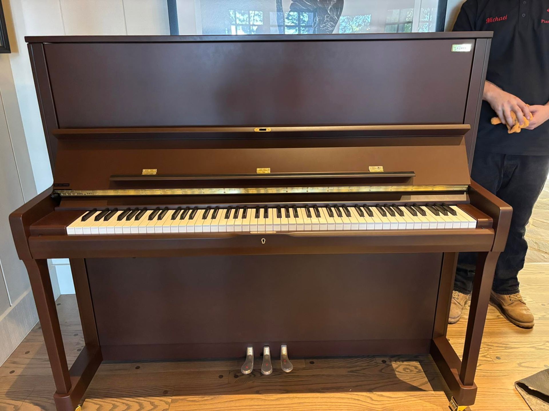 Brown upright piano in a room, with a person standing partially visible beside it.