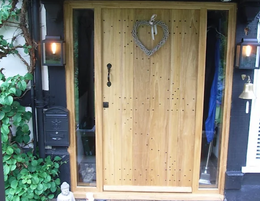 A wooden front door with a heart-shaped wreath, flanked by glass panels, black lanterns, and a mailbox on a vine-covered wall.