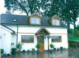 A two-story white house with a dark roof, wooden accents, two dormer windows, and a paved front patio with potted plants.