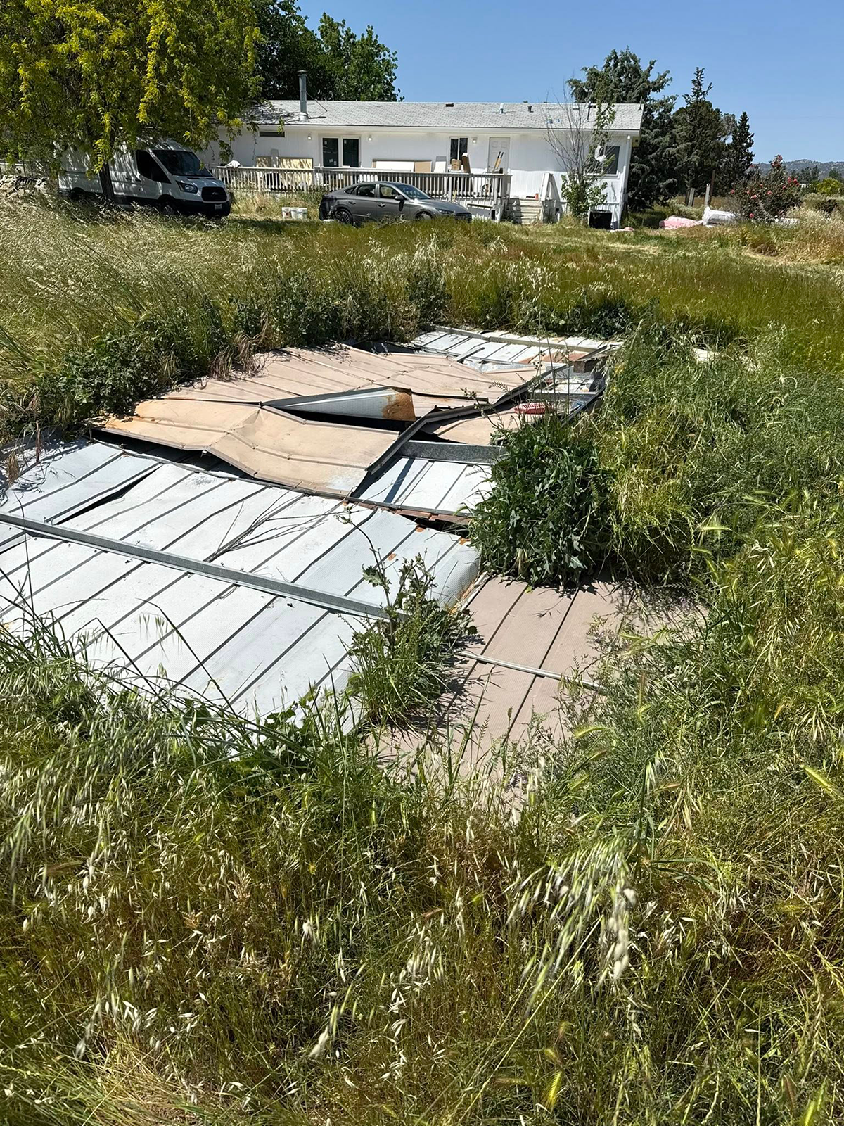 Overgrown lot with debris in front of a white house on a sunny day.