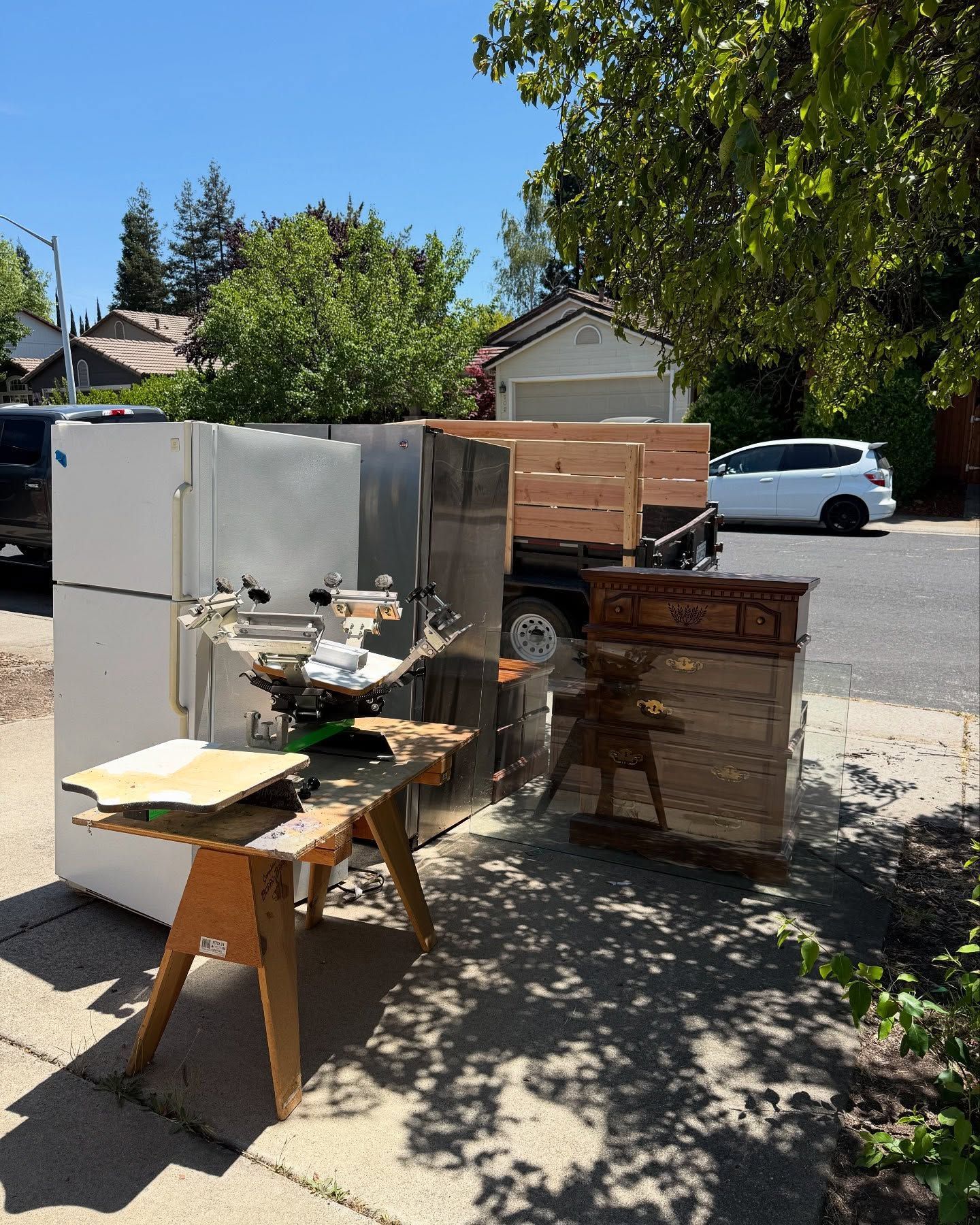 Several large appliances, a trailer, and tools on a sunny residential street.