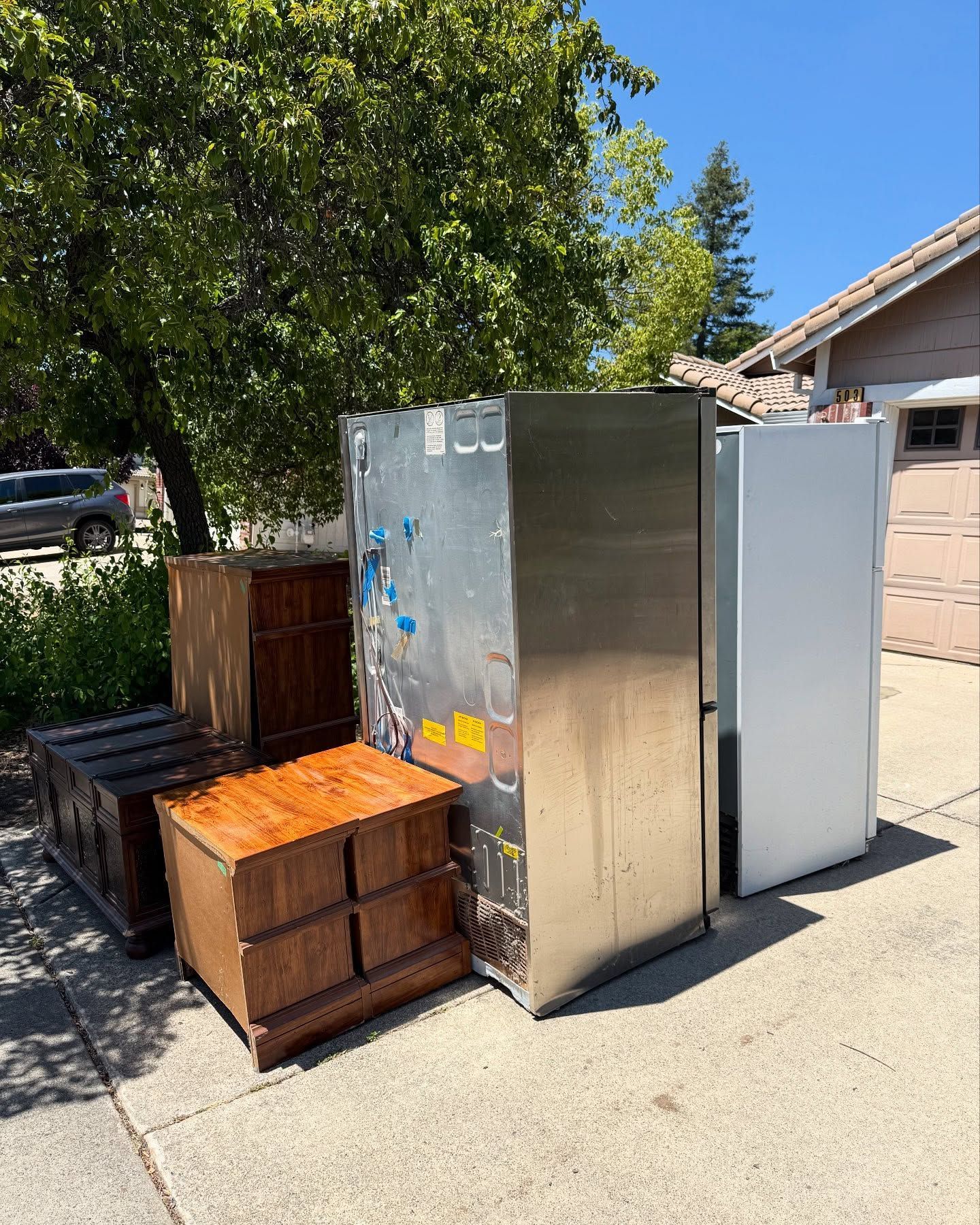 Pile of discarded furniture and appliances on a driveway; includes dressers, a refrigerator, and a freezer.