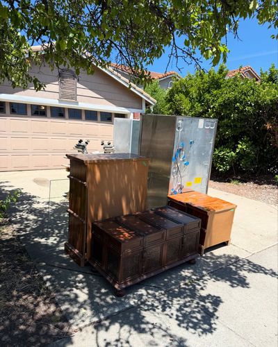 Discarded furniture and metal sheets on a sidewalk in front of a house with a closed garage door, under tree shadows.