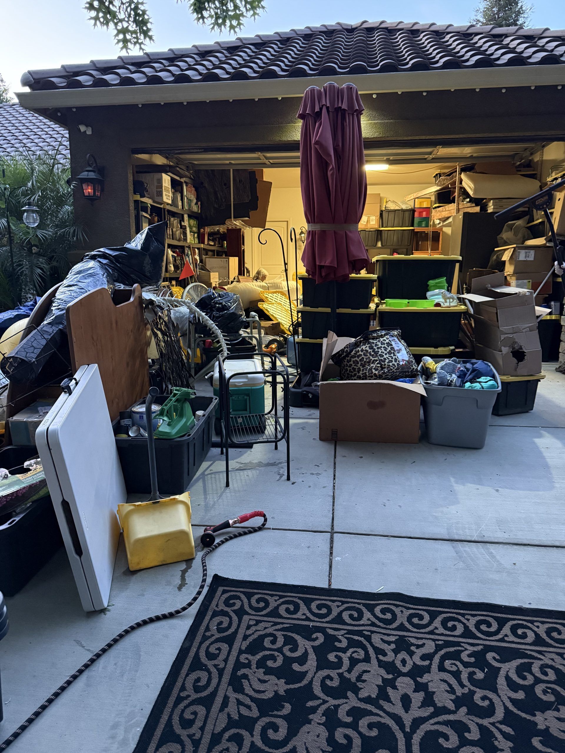 Garage sale with items scattered on driveway. Boxes, furniture, and an umbrella are visible.
