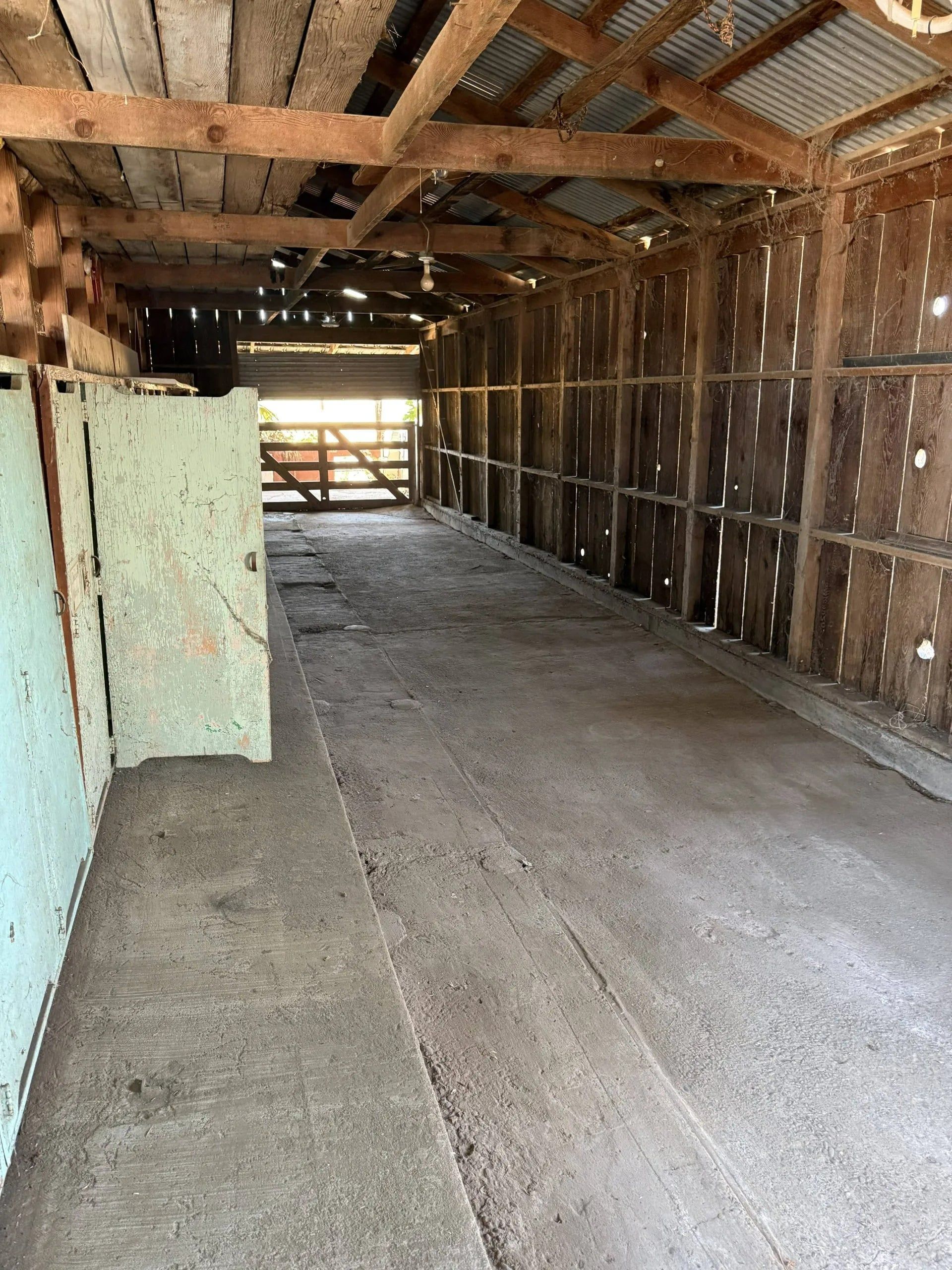 Inside of an old barn with wooden beams, a dirt floor, and open gate.