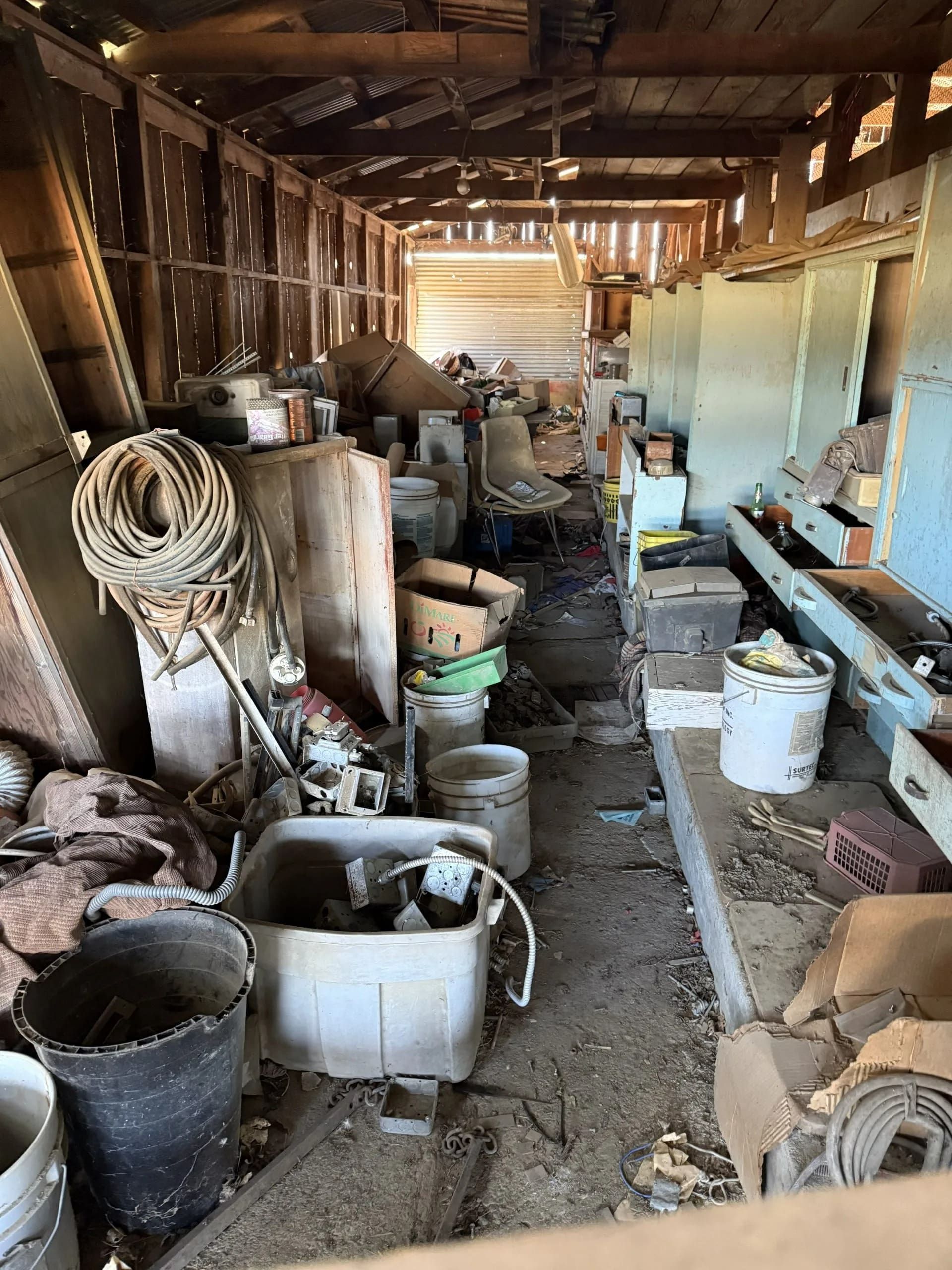 Interior of a cluttered shed filled with tools, buckets, and debris. Wooden structure with a central aisle.