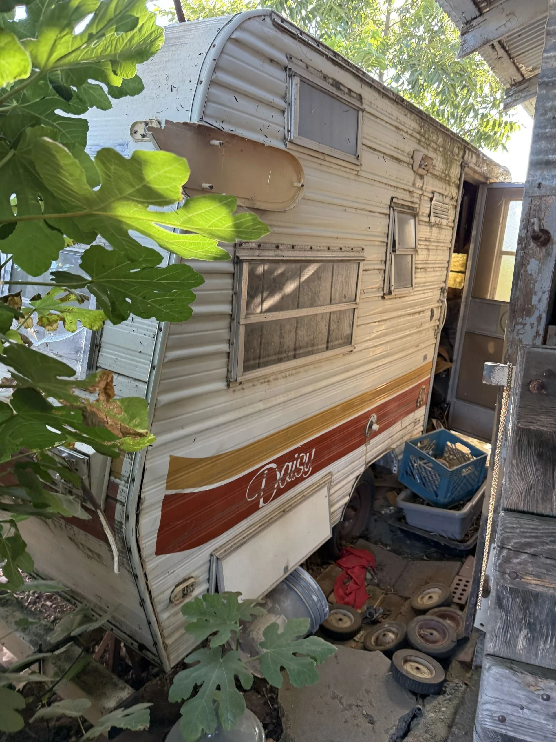 Old, weathered camper trailer in disrepair, leaning, with faded red and orange stripes.