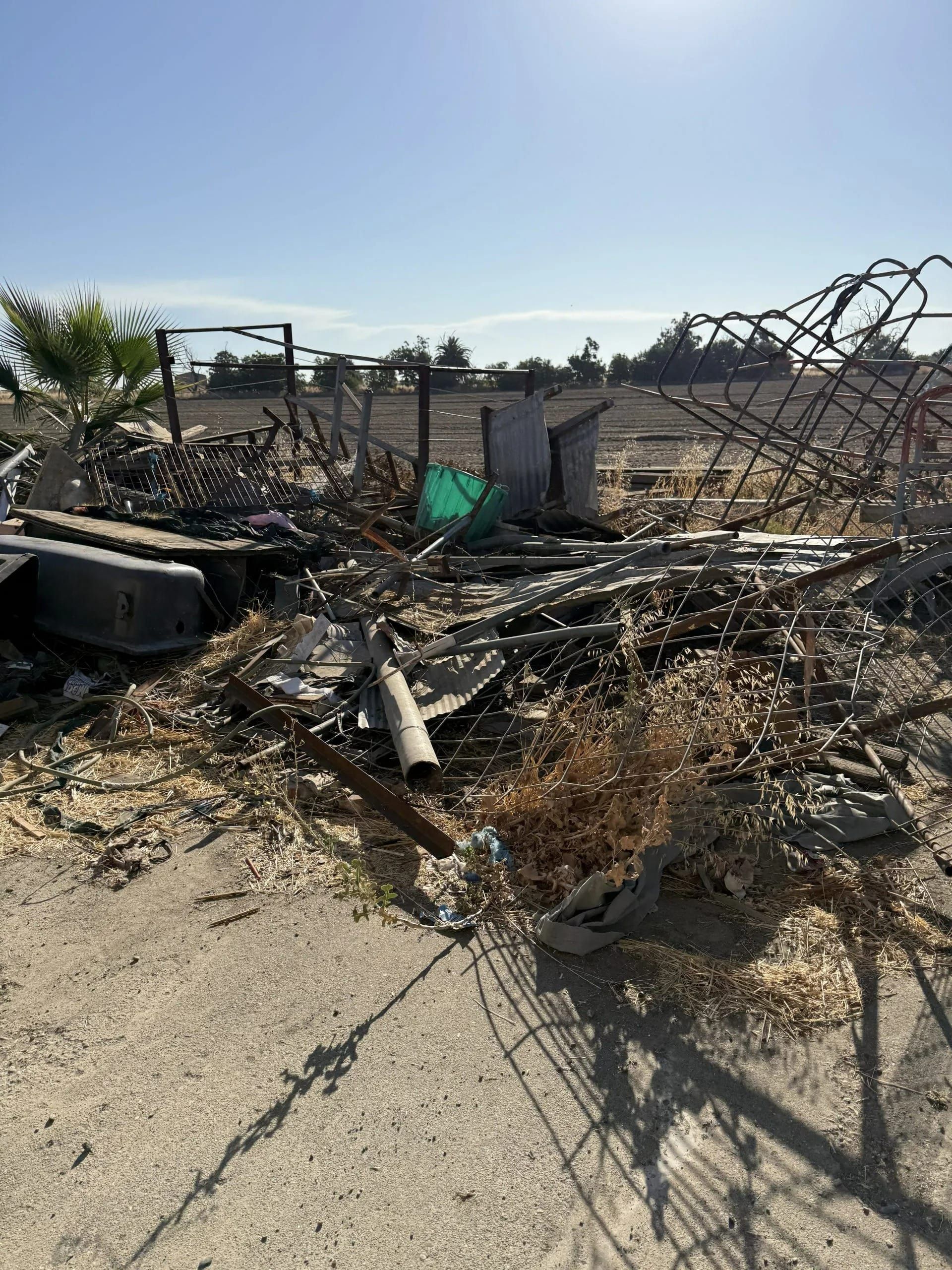 Debris pile with metal, dried grass, and a barbed wire fence, set against a clear blue sky.