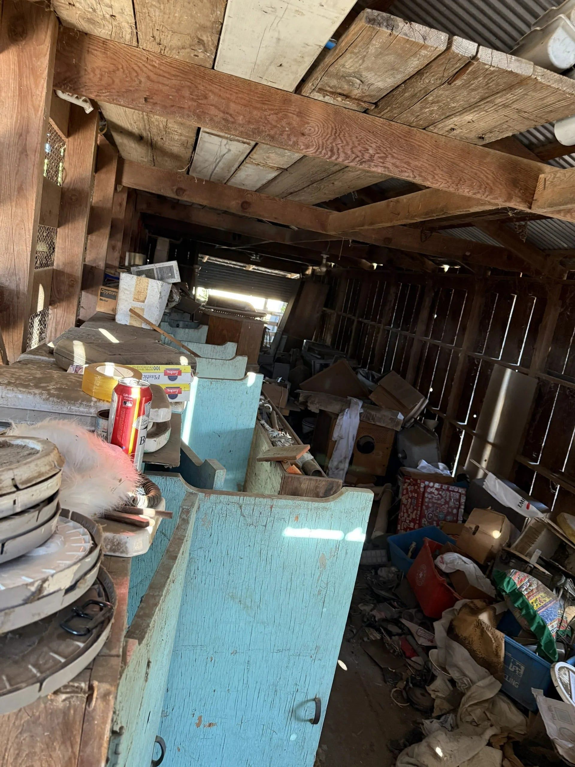 Cluttered interior with wooden beams, boxes, and various objects, in a poorly lit, dusty storage area.