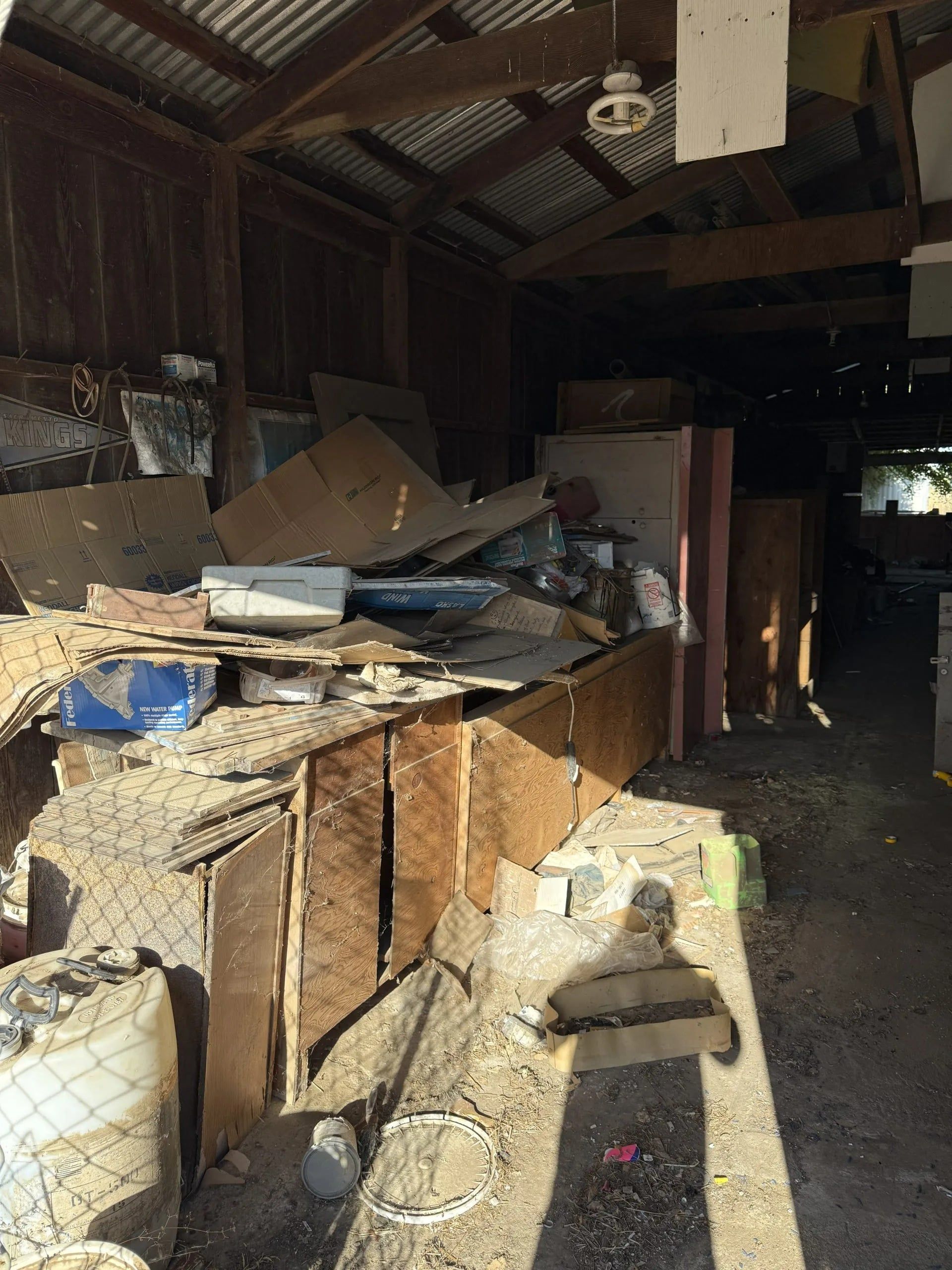 A cluttered shed interior filled with piles of junk, including wood and metal, under a corrugated roof.