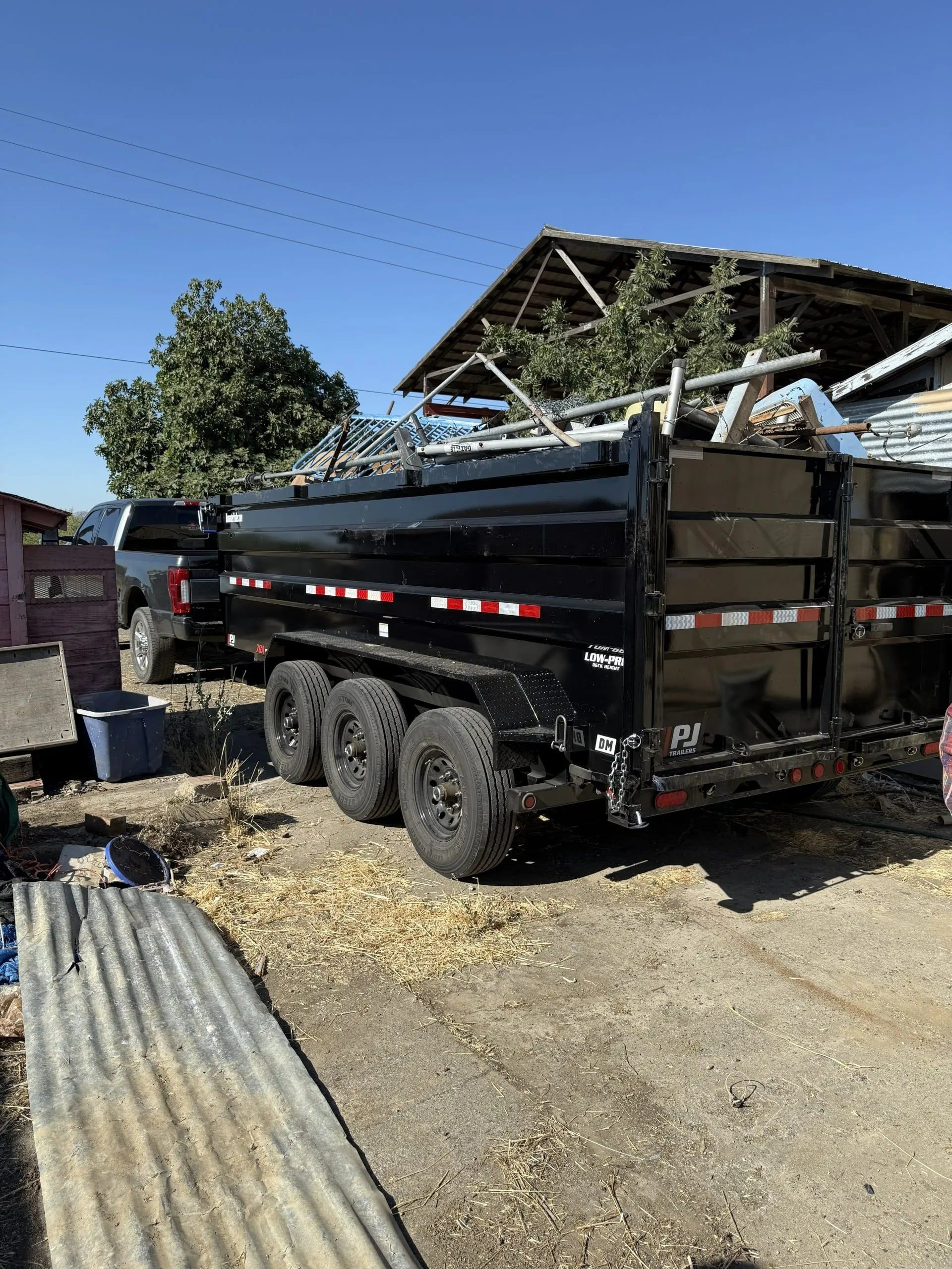 Black dump trailer hitched to a truck, parked outdoors near a weathered building on a sunny day.