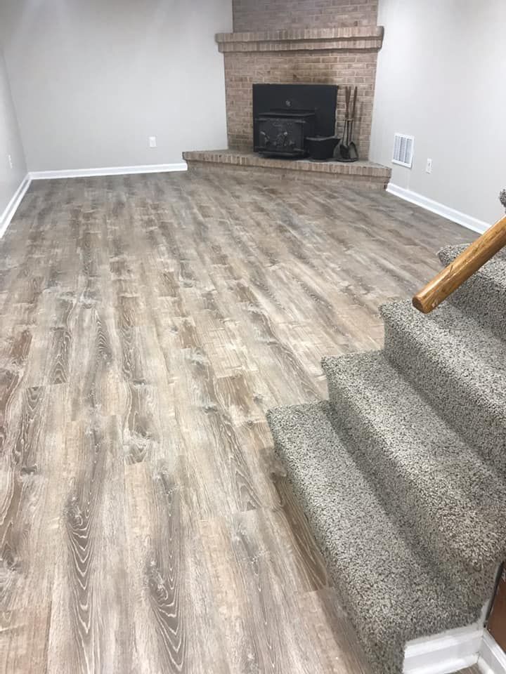 Basement with wood-look flooring and a raised brick fireplace; carpeted stairs on the right.