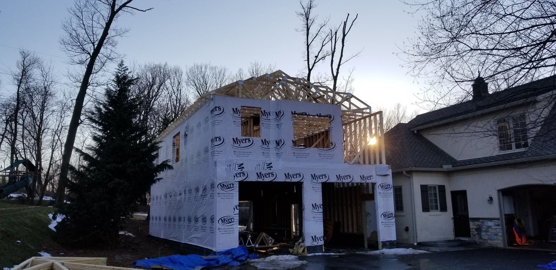 A two-story building under construction; unfinished frame and garage doors. Daytime, with trees in the background.