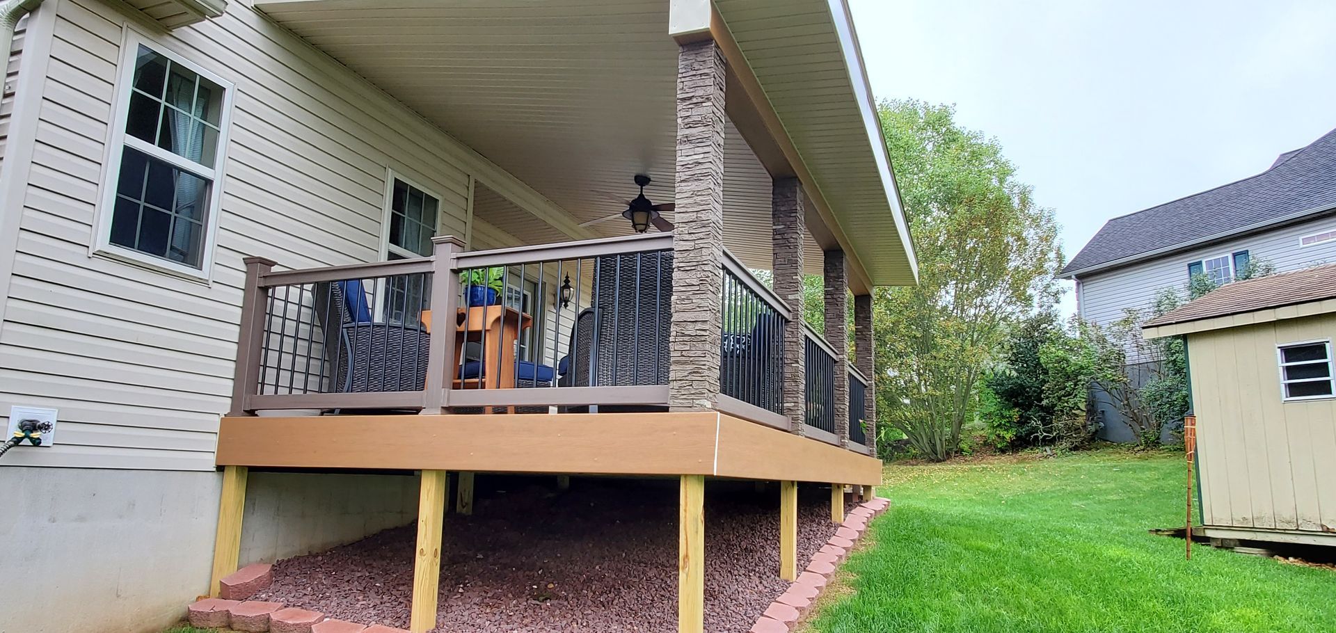 A covered deck with stone columns and a view of a grassy yard, a small shed, and a house.