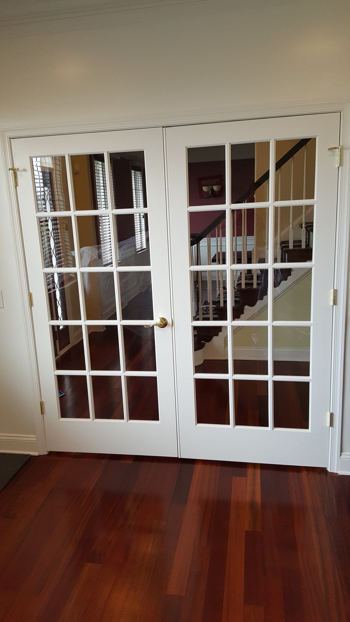 White French doors with multiple glass panes, reflecting a hallway with a staircase and a dark red wall.