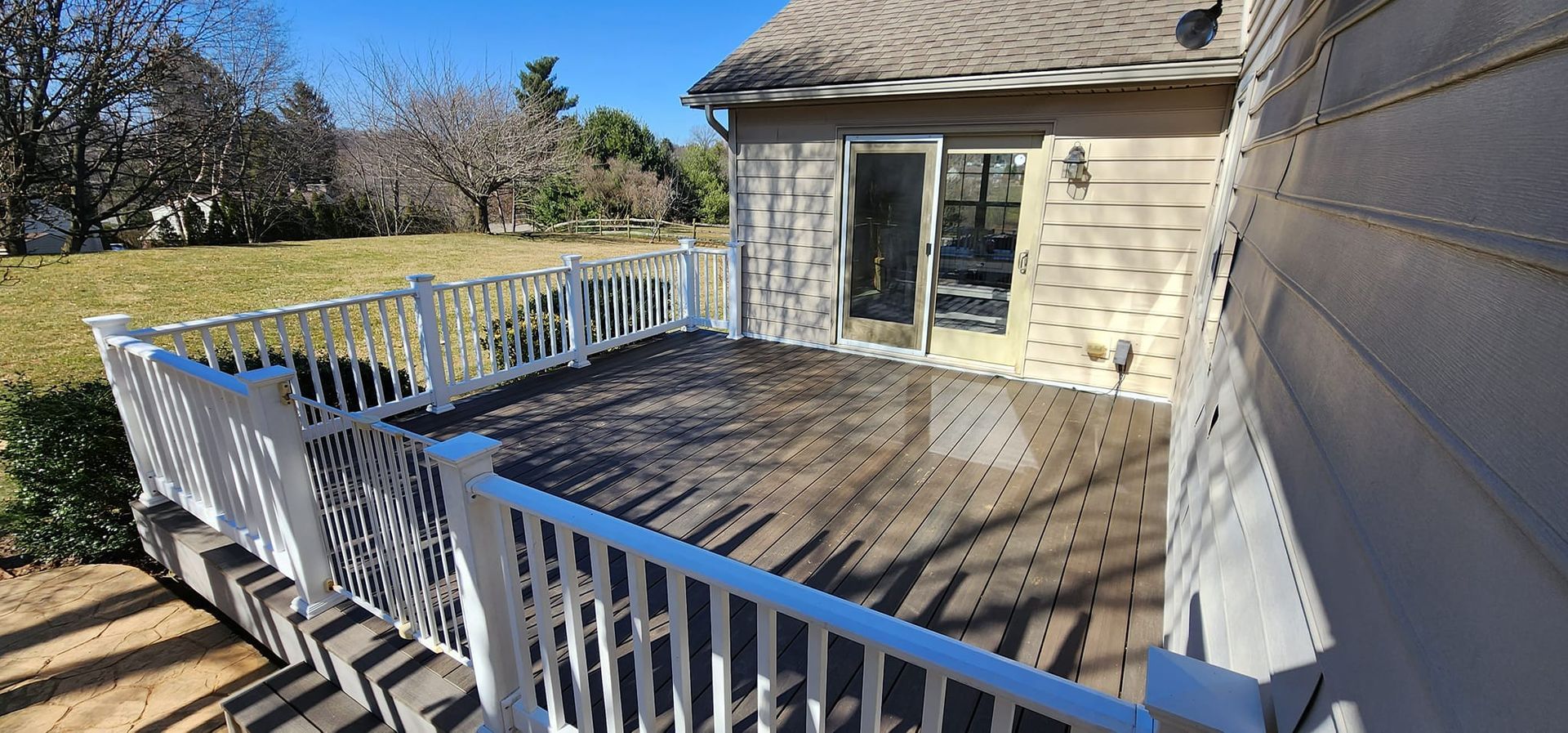 Wooden deck with white railing next to a house, with grass and trees in the background.