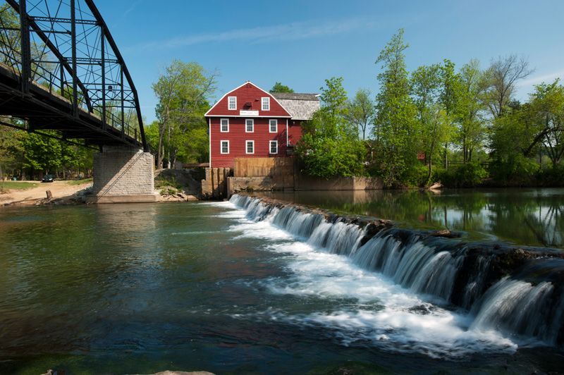 A red barn is sitting on the shore of a river next to a waterfall.