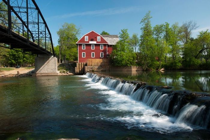 A red barn is sitting on the shore of a river next to a waterfall.