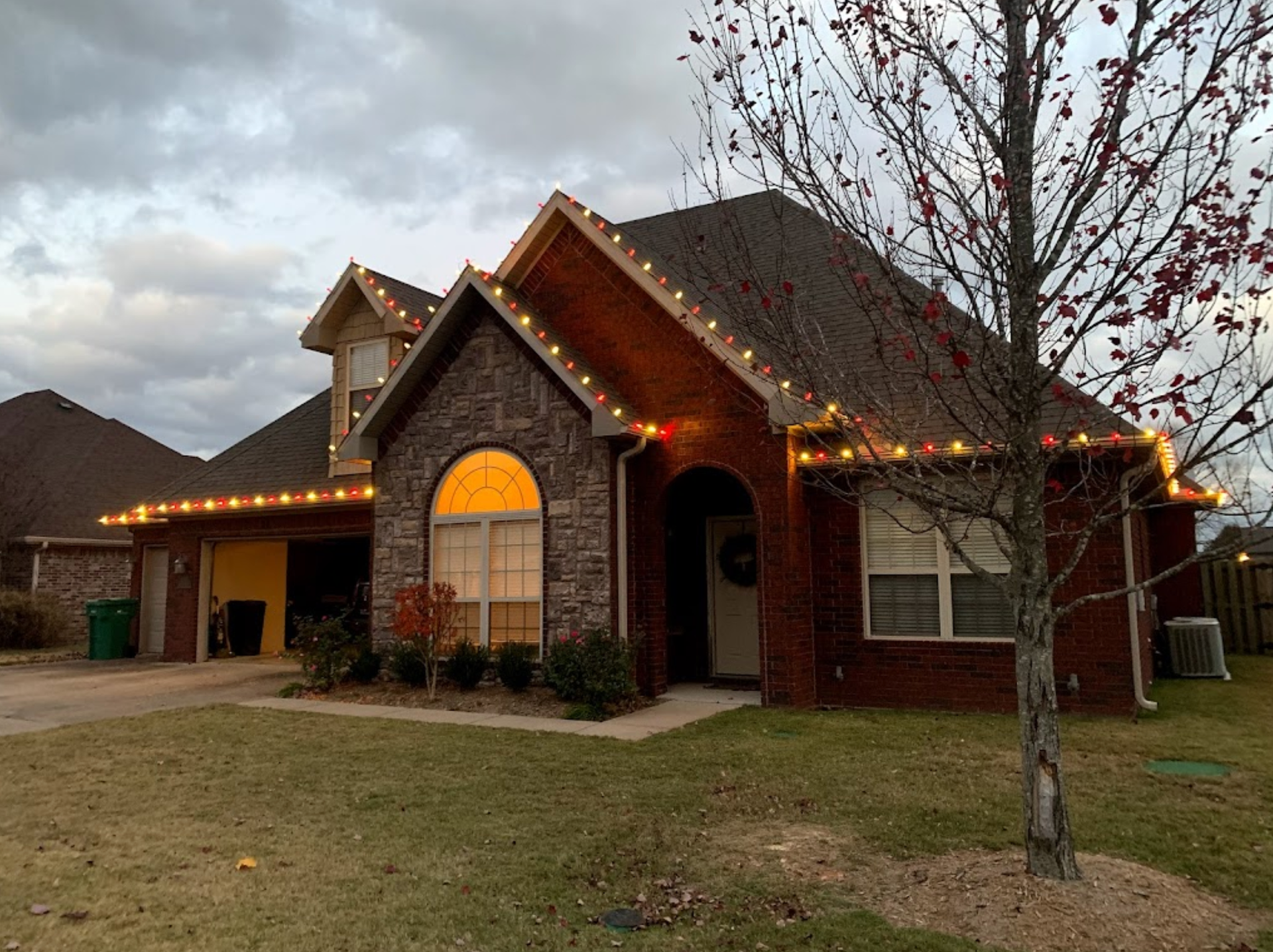 A brick house with christmas lights on the roof and a tree in front of it.