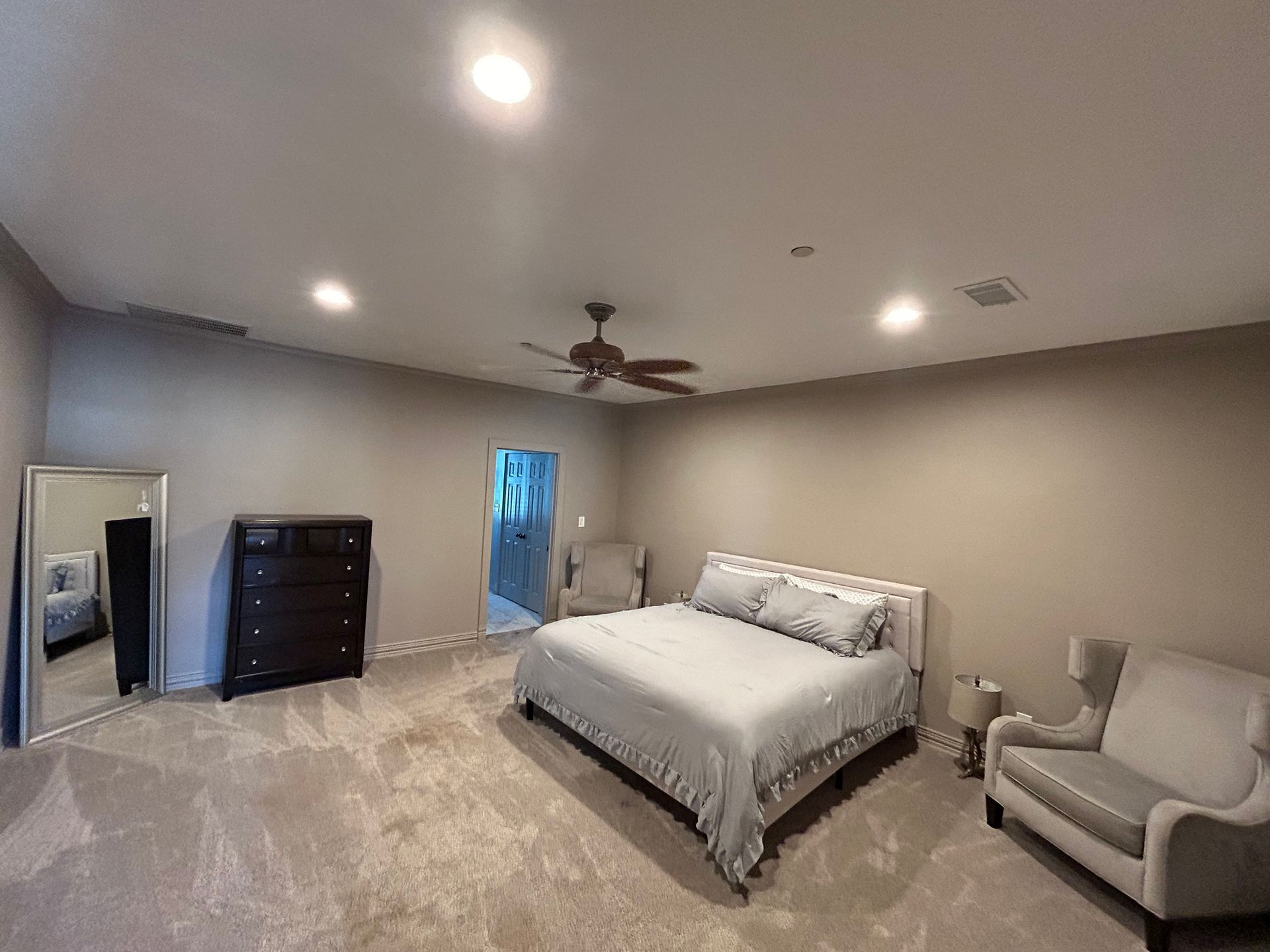 A bedroom with a large bed , dresser , chair , mirror and ceiling fan.