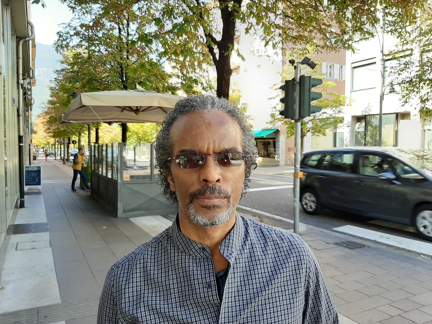 Man with graying hair and sunglasses stands on a sidewalk in front of a building; a car and streetlights are visible.
