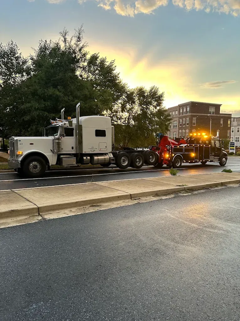 White semi-truck being towed by a tow truck on a wet road during a sunset, buildings and trees in the background.
