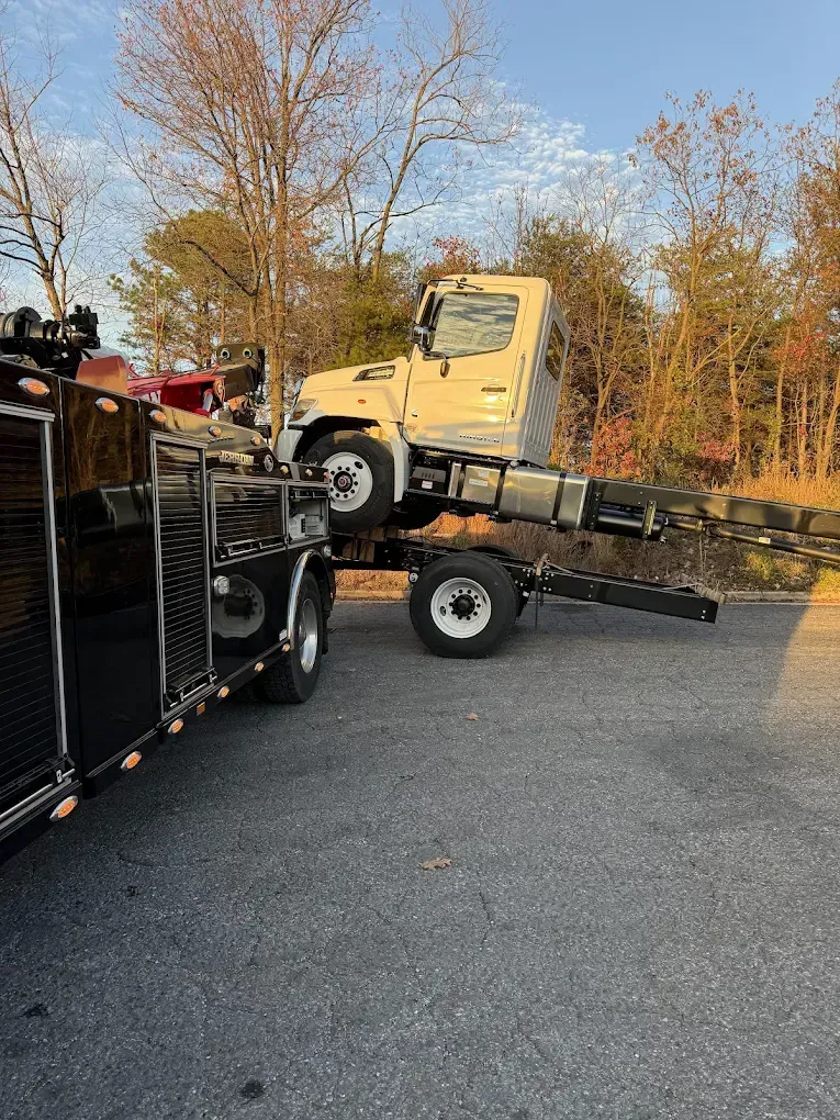 A tow truck is loading a tan cab-over truck on an asphalt lot. Trees in background.