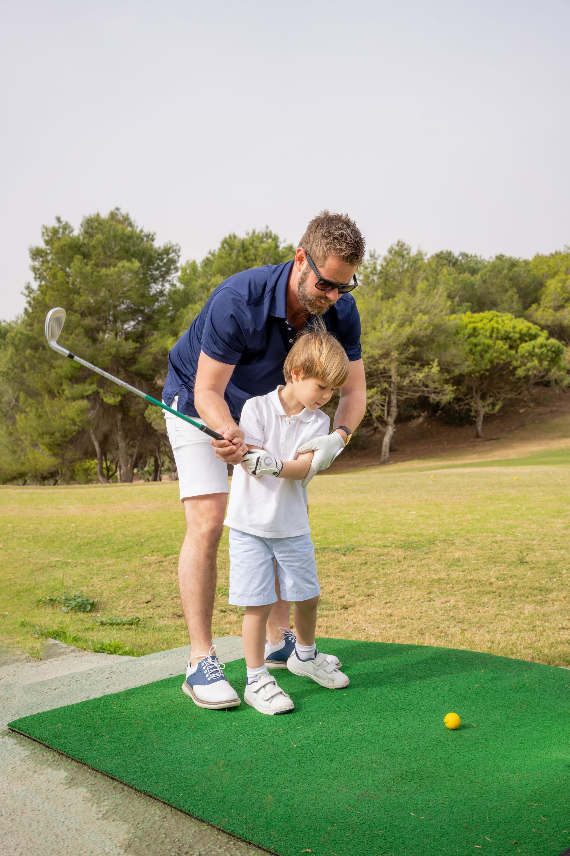 Man teaching a young boy how to golf, outdoors on green turf. Both are wearing golf attire.