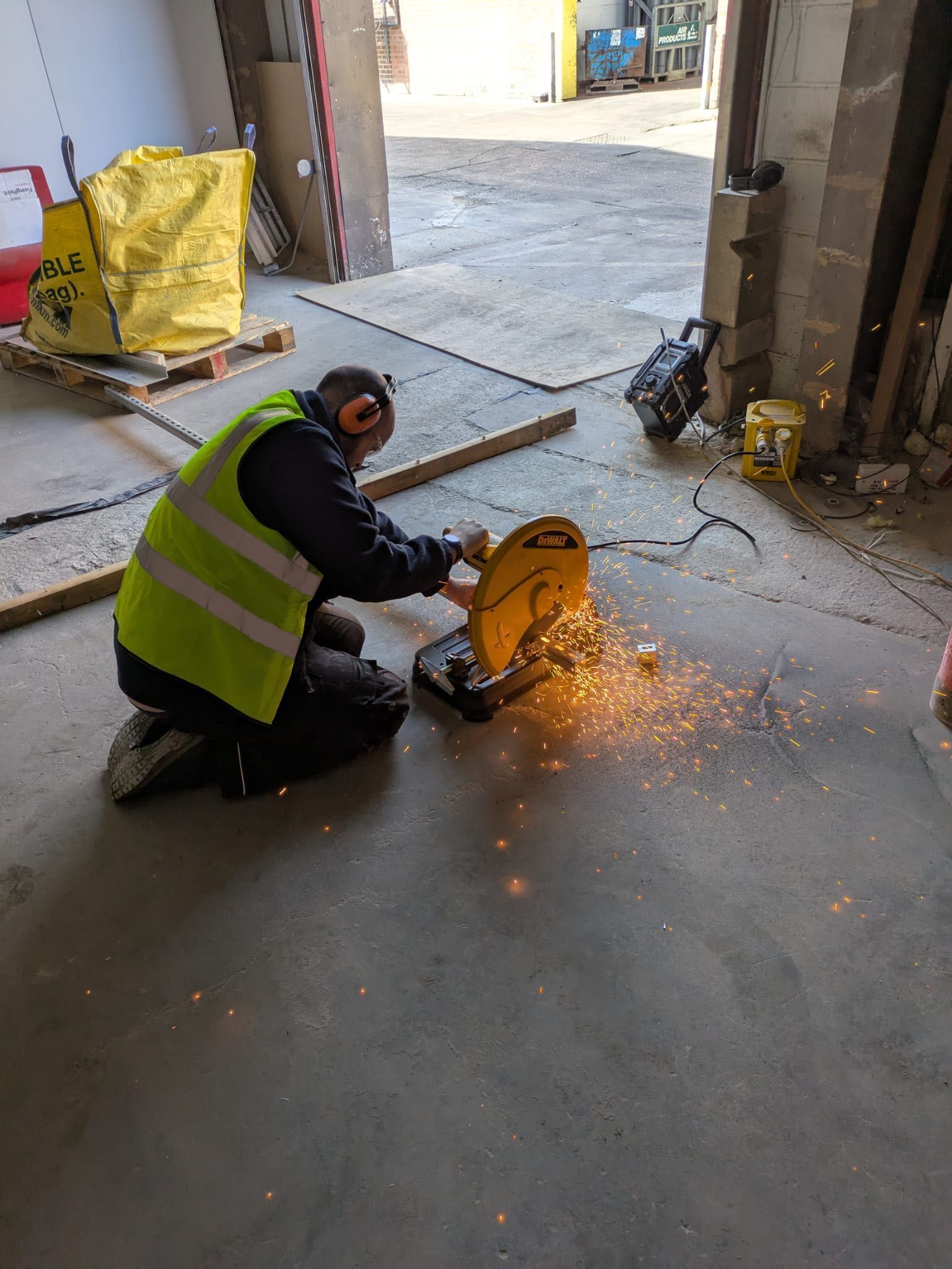 Worker in yellow safety vest cuts metal with a saw, sparks fly in a construction setting.