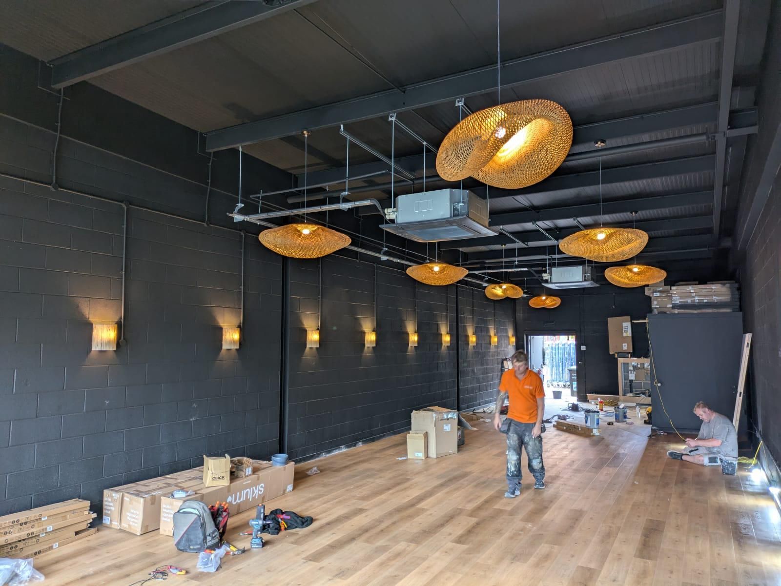 Interior of a room under construction with dark walls, wooden floor, decorative lights, and workers.