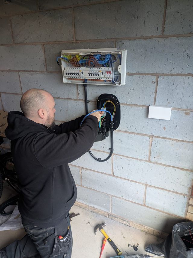 Electrician installing an EV charger on a cinder block wall, wiring connected to a breaker box.
