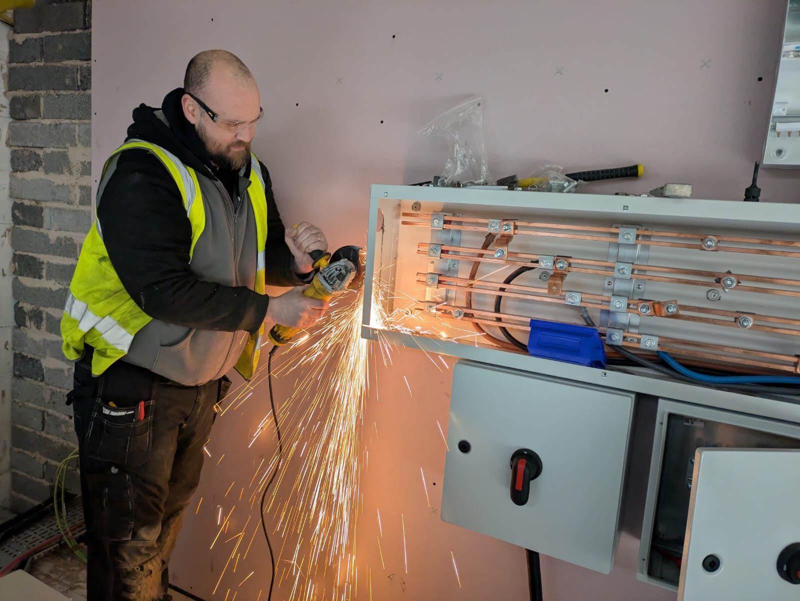 Electrician using a grinder, creating sparks, cutting a metal electrical panel mounted on a wall.