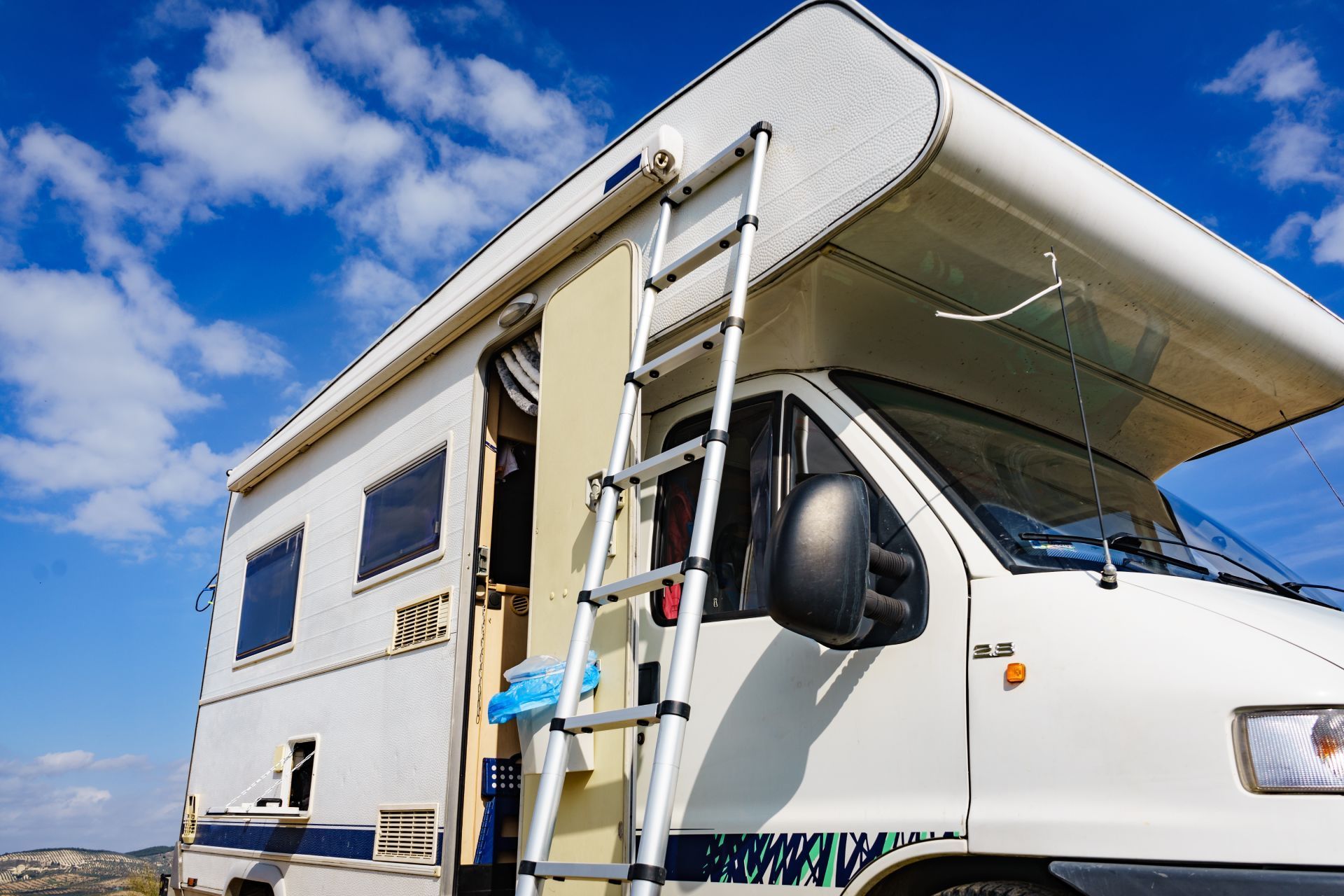 White RV with a ladder extended, parked against a blue sky with clouds.