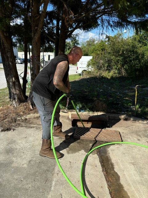 Man in black tank top and work boots washes a drain with a green hose outdoors.