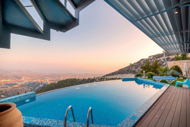 An infinity pool overlooking a city at sunset from a terrace with wooden decking and modern overhead shading.