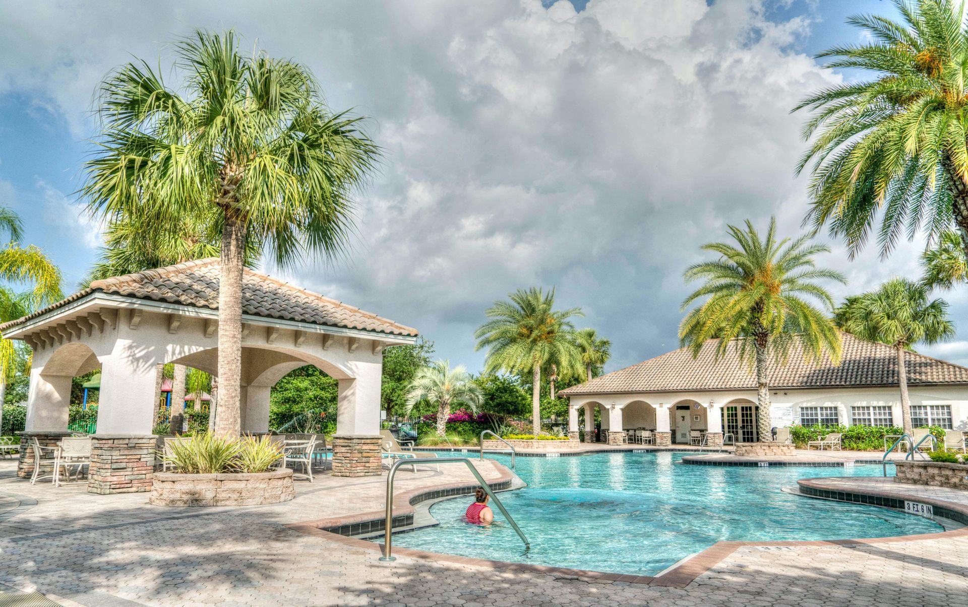 A resort-style swimming pool with palm trees, a stone gazebo, and a clubhouse under a cloudy sky.