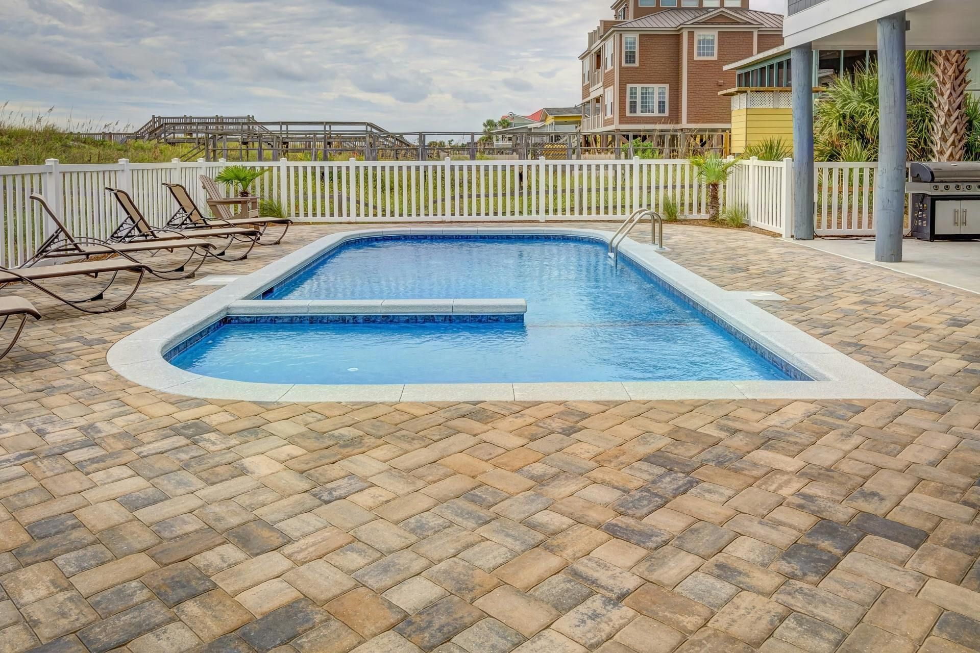 A rectangular pool with a built-in step, surrounded by tan stone pavers and a white fence in a coastal setting.