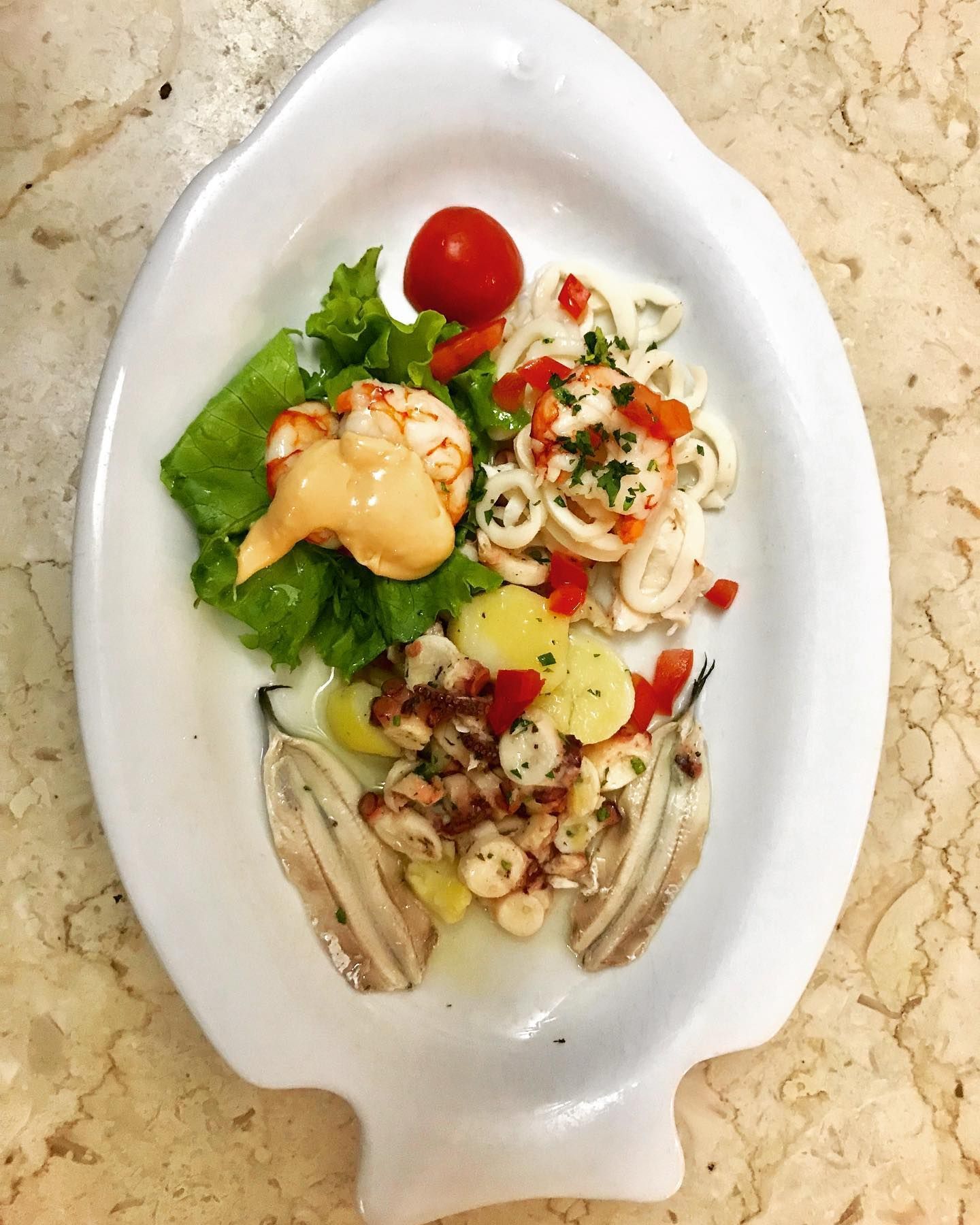 Un antipasto di pesce con gamberi, calamari, polpo, patate e acciughe servito su un piatto a forma di pesce.