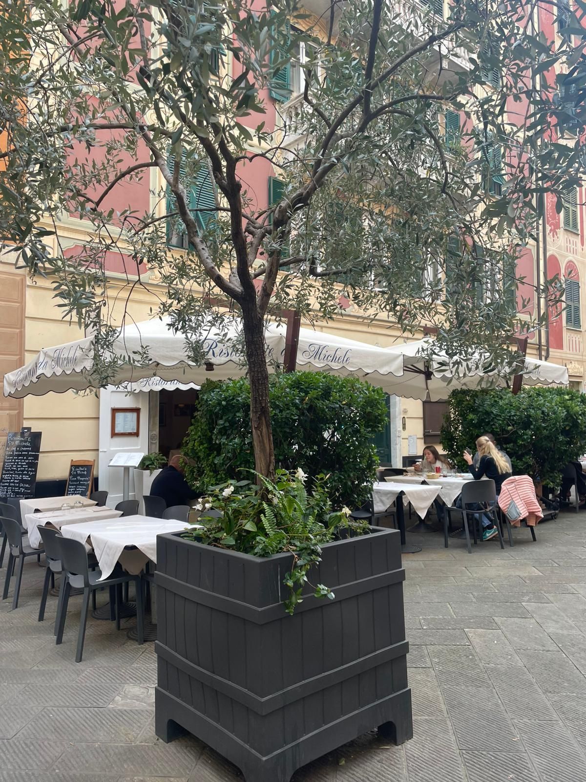 La terrazza di un ristorante affacciato sulla strada, con tavoli e sedie apparecchiati con tovaglie bianche e una grande fioriera centrale sotto la tettoia di un edificio.