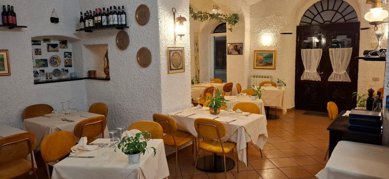 Interno di un accogliente ristorante con pareti bianche, pavimento in cotto, tavoli apparecchiati con sedie gialle e vino sugli scaffali.