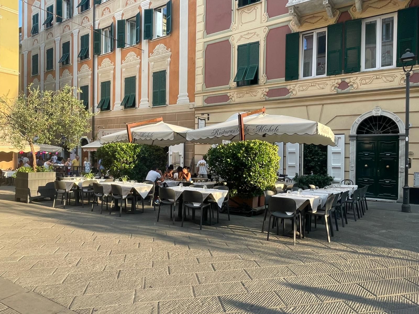 Tavolini e sedie all'aperto in un caffè, riparati da grandi ombrelloni bianchi, su una piazza lastricata di ciottoli di fronte a edifici storici.